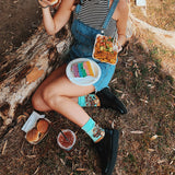 Person sitting outdoors with food, wearing denim overalls and colorful socks. thumbnail