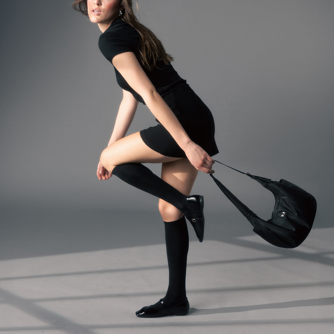 Woman in black outfit with knee-high socks and a black handbag on a gray background