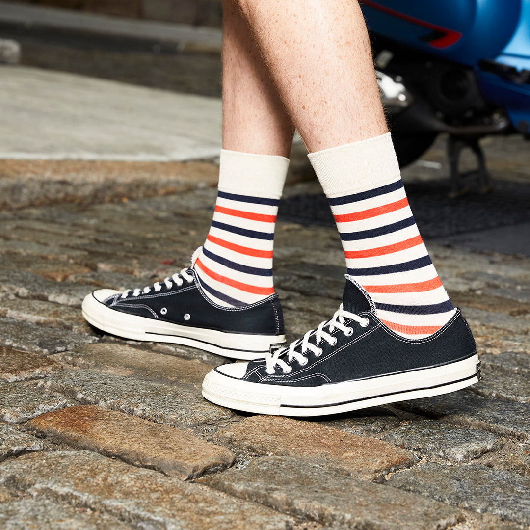 A person wearing navy low-top canvas sneakers with white stripes and cream, navy, and orange striped socks, standing on a cobblestone street.