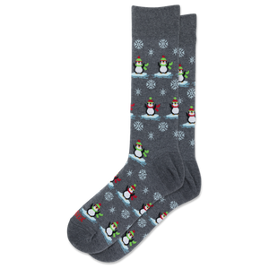 A pair of dark gray crew socks featuring a repeating pattern of penguins wearing red and green hats and scarves, standing on icy blue platforms with white snowflakes.