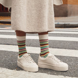 A person standing on a crosswalk wearing cream platform sneakers, a beige knit skirt, and white crew socks with multicolored horizontal stripes.