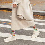 A person walking across a crosswalk wearing a long cream knit dress, a light beige cardigan, colorful striped socks, and white platform sneakers.