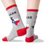 A pair of crew socks worn on feet, shown against a plain white background. The socks are heather gray with red cuffs, heels, and toes, and feature a graphic of the state of Texas with the word 'TEXAS' above it.