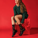 A woman sits on a large red gift box against a solid red studio background, wearing a dark green cable-knit sweater dress, black leather shoes, and black crew socks with a colorful Christmas tree pattern.