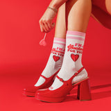 A person seated against a bright red studio background wearing white socks with pink stripes and red text that reads “I’m a Sucker for You,” paired with glossy red Mary Jane-style heels with block heels and ankle straps.