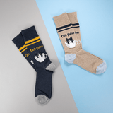 Two pairs of crew socks displayed on a split gray and light blue background. One pair is navy blue with yellow stripes and a cat-in-a-teacup graphic, and the other pair is heather beige with dark navy stripes and a matching cat-in-a-teacup graphic. thumbnail