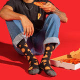 A person sitting on a bright red floor wearing black socks with a repeating pizza slice pattern, holding a slice of pizza. A pizza box is open on the floor beside them. thumbnail