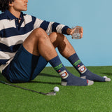 A man sits on artificial grass wearing navy and cream striped polo shorts and dark blue crew socks with a golf-themed graphic. He holds a glass, with a golf club and golf ball nearby against a solid light blue background. thumbnail
