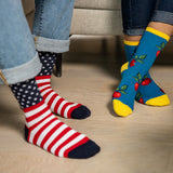 Two people wearing colorful novelty crew socks, one with red and white stripes and a navy star pattern, and the other with a blue cherry print and yellow toes, standing on a light wood floor.