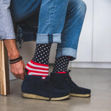 A person wearing blue jeans and dark navy suede boots, with one hand adjusting patriotic socks that feature a white star pattern on a black background and red-and-white stripes near the top.