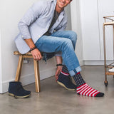 A man sitting on a wooden stool wearing blue jeans, a light blue shirt, and navy blue boots, while trying on a pair of socks with an American flag pattern. The socks feature red and white stripes on one leg and a navy blue background with white stars on the other, with red cuffs.