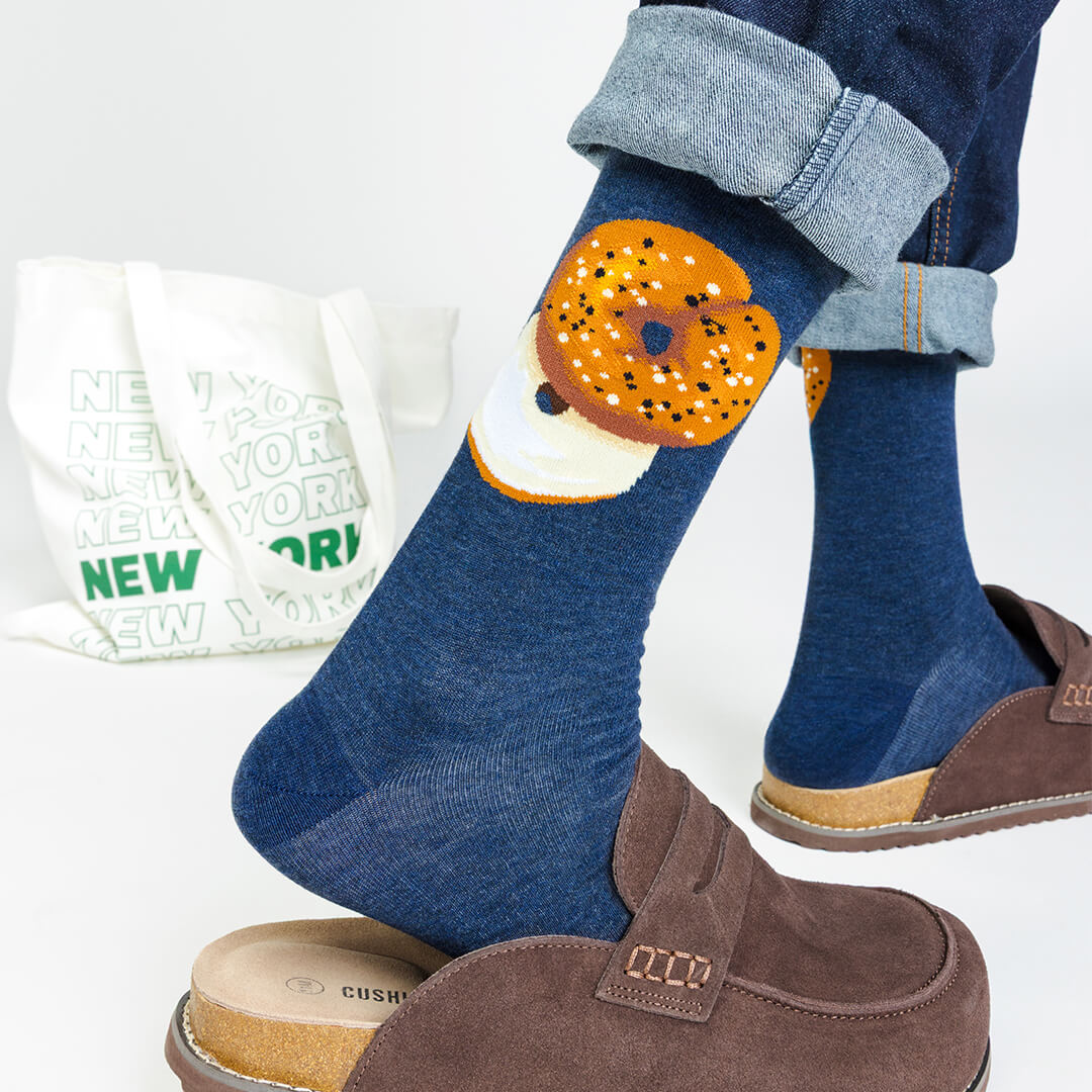 Person wearing blue socks with a bagel design, paired with brown shoes and a white bag with 'New York' text.