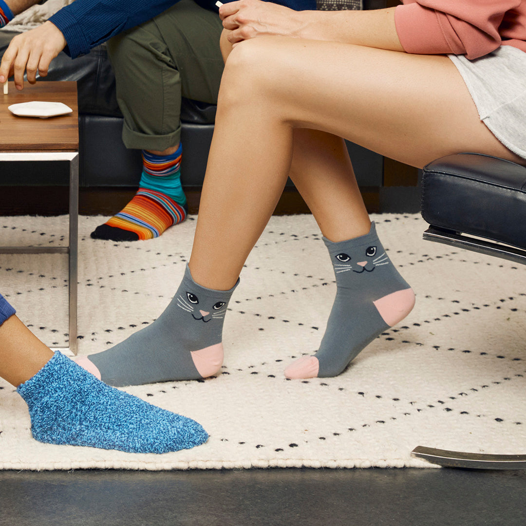 Gray ankle socks with a cat face design worn by a person sitting on a couch in a living room setting, alongside a blue knit slipper and another person's legs.