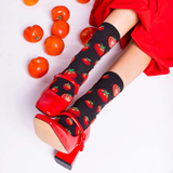 Person wearing red high heels and black socks with tomato pattern, surrounded by tomatoes on a white background
