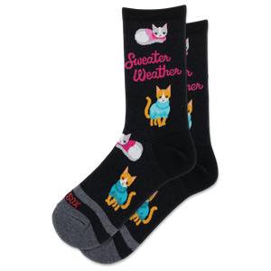 A pair of black crew socks with a cozy pattern of orange and white kittens wearing blue and pink sweaters, alongside the pink text 'Sweater Weather'. The toes and heels are gray, and the word 'SOX' is printed on the toe.