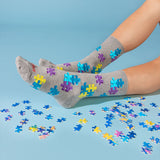 A pair of gray crew socks with colorful jigsaw puzzle-piece patterns, worn on feet with scattered puzzle pieces on a light blue background.