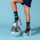 A person wearing black crew socks with a colorful space-themed graphic and silver high heels, posed with one foot on a mirrored disco ball against a light blue studio background.