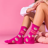 A person seated against a pink studio background wearing bright pink crew socks printed with cupcake illustrations and the phrase “DEM BUNS DOUGH.”. thumbnail