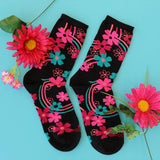 A pair of black crew socks decorated with vibrant pink, teal, and orange floral patterns, arranged on a light blue background with decorative pink flowers.