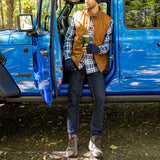 Person standing next to a blue Jeep in a forested area, holding a drink.