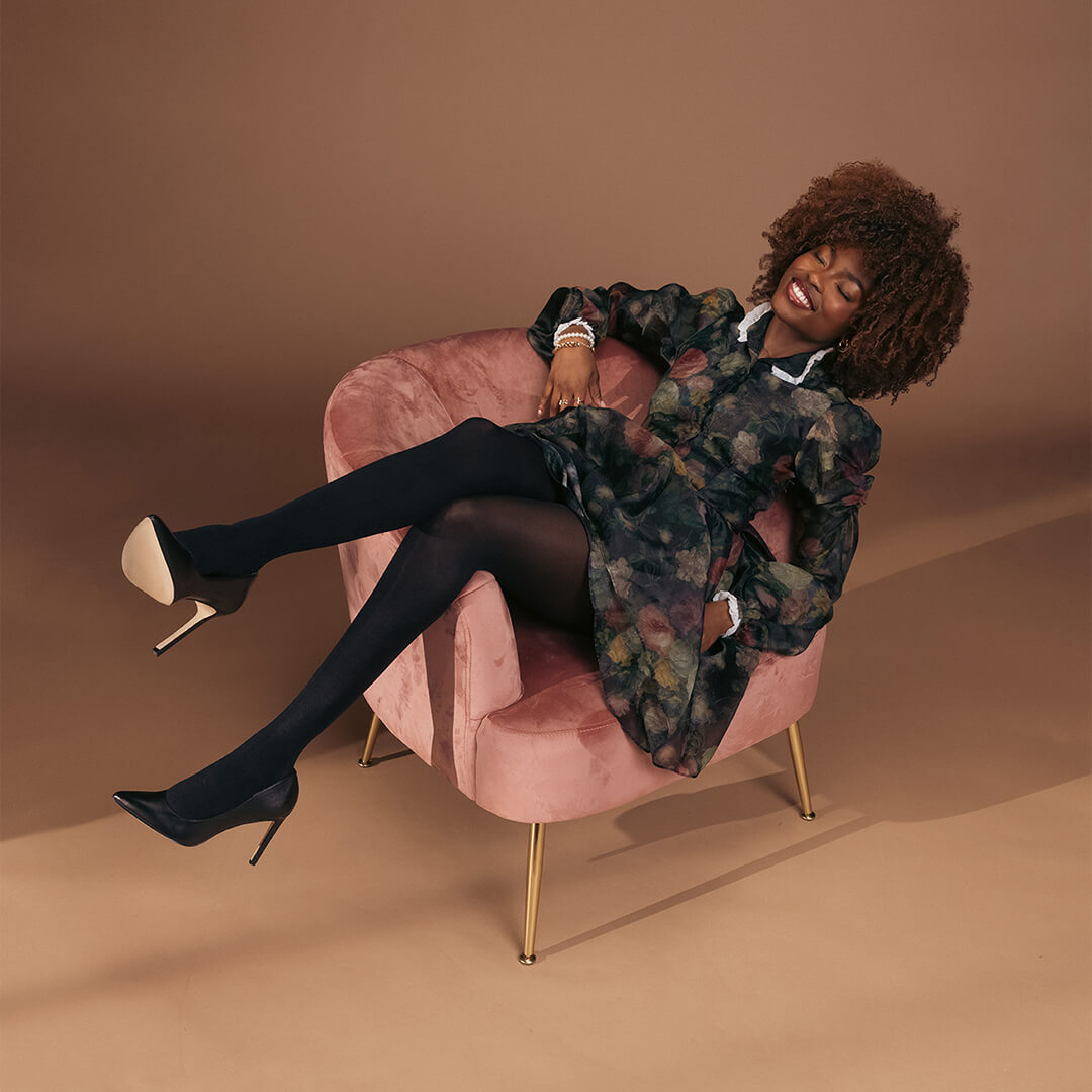 Woman sitting on a pink chair wearing a floral dress with black tights and heels on a brown background