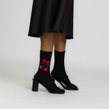 A person wearing black mid-heel sandals and black crew socks with a red cherry and bow graphic, shown from the knees down against a light gray studio background.