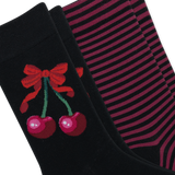 Two pairs of black crew socks arranged diagonally. The front sock features a red bow and two pink cherries with green stems, while the back pair has thin horizontal pink stripes.