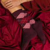 A person wearing midi-crew argyle socks in shades of burgundy, dark red, and pink, resting on a draped burgundy satin fabric. thumbnail