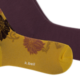 A close-up of two socks from k.bell, featuring a metallic gold sock with a floral pattern and white brand text, layered over a solid dark plum purple sock, on a white background.