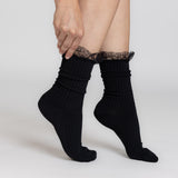 A person wearing black ribbed ankle socks with a black lace ruffle at the top, shown against a light gray studio background. thumbnail
