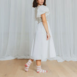 Woman wearing a white dress with a patterned pink and white striped socks standing in front of white curtains.