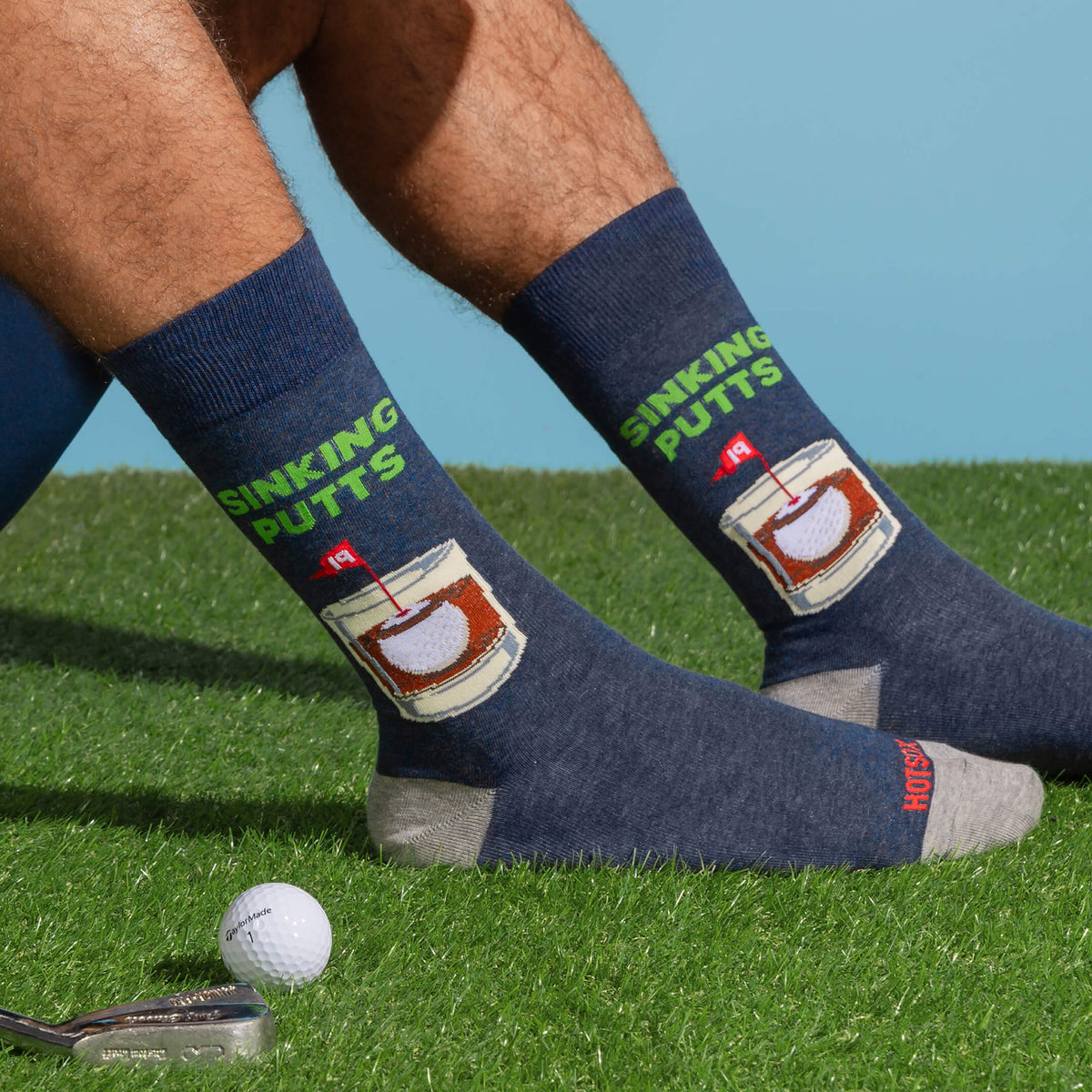 A person sitting on a golf course grass wearing dark navy crew socks with a green 'SINKING PUTTS' text design and a graphic of a bourbon glass. A golf ball and putter are visible on the grass.