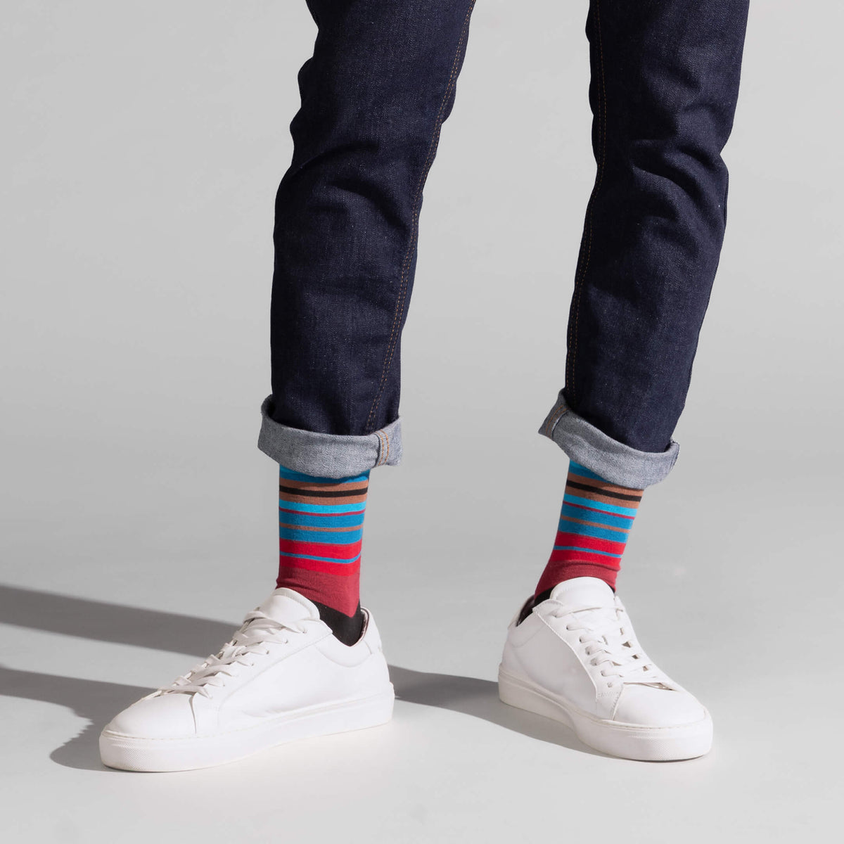 A person wearing dark blue jeans with cuffed hems, white sneakers, and colorful striped socks, standing against a light gray studio background.