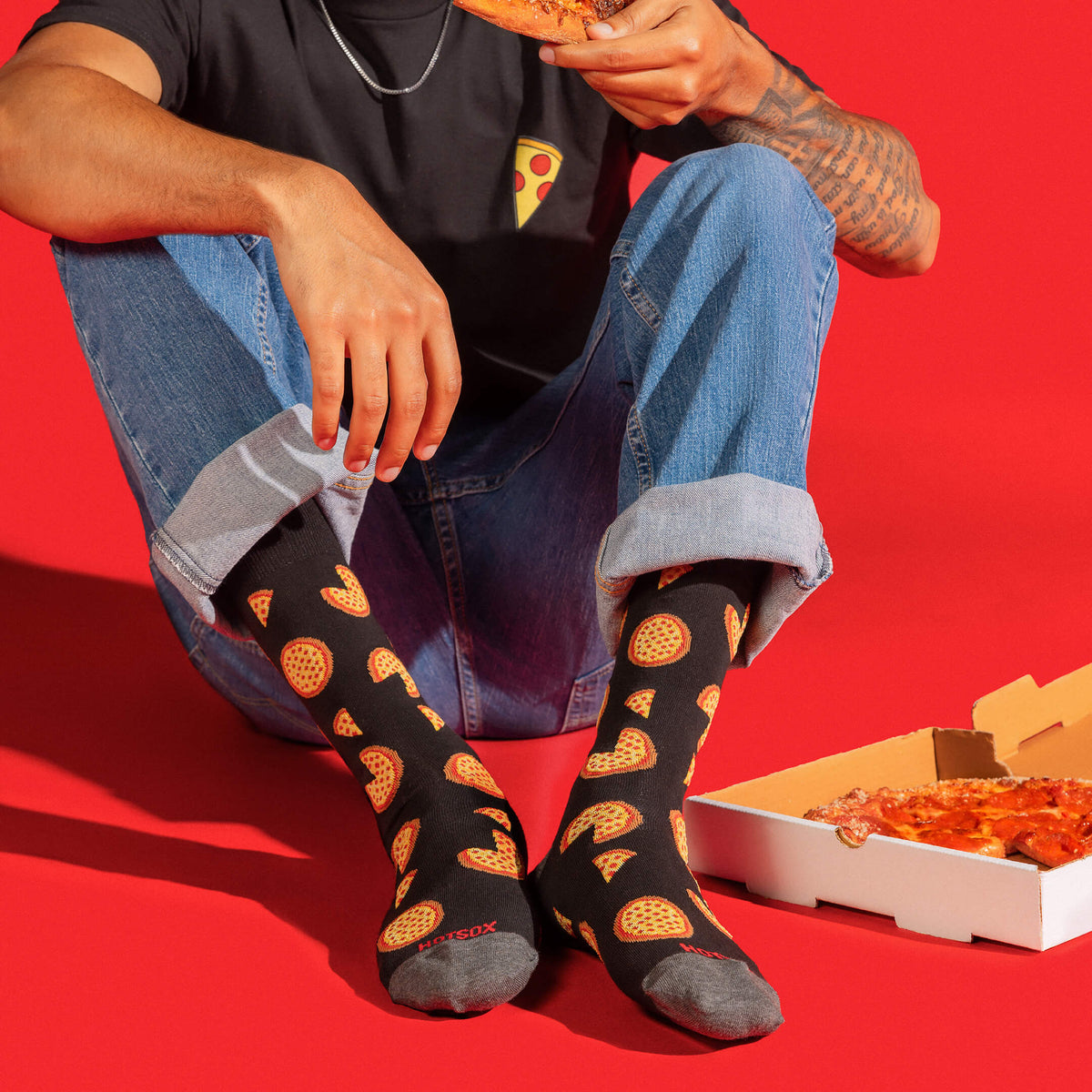 A person sitting on a red floor wearing black crew socks with a repeating pizza slice pattern, blue jeans, and a black t-shirt, while holding a slice of pizza next to an open pizza box.