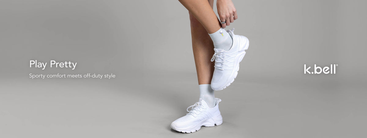 A person wearing white K. Bell embroidered golf anklet socks, shown against a warm gray studio background with product text on the left that says, 'Play Pretty - Sporty comfort meets off-duty style'