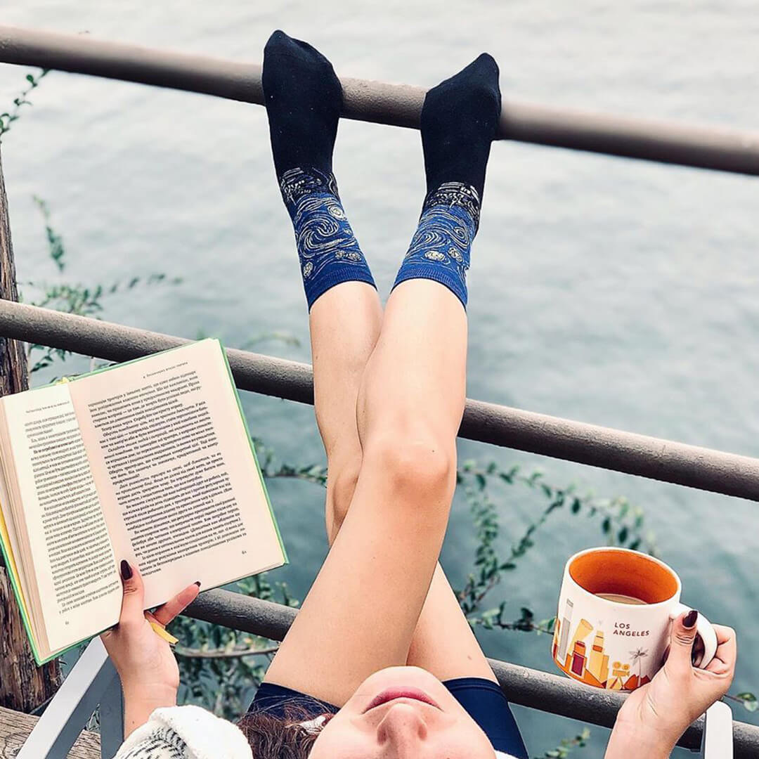 Person sitting by a body of water with legs crossed, wearing blue socks and holding a book and mug.