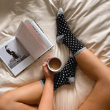 Woman holding a coffee cup, sitting on a bed wearing HOTSOX black and white polka dot socks while reading a book.