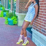Woman in a light blue dress and yellow high-heeled shoes leaning against a brick wall with plants in the background. thumbnail