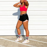 Person wearing a red sports bra and black shorts with 'Under Armour' branding, standing against a white wall. thumbnail