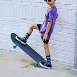 Child in purple shirt and black shorts standing on a skateboard against a white brick wall.