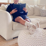 Person sitting on a couch reading a book with their feet propped up on a poof wearing HOTSOX coffee cup socks thumbnail