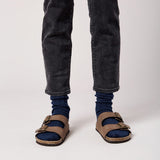 Person wearing blue socks, dark jeans, and brown sandals on a white background
