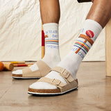 Person wearing white socks with colorful text and designs, paired with beige sandals on a wooden floor. thumbnail
