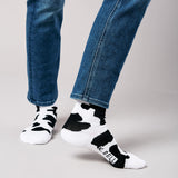 Person wearing black and white cow print socks with 'K-Bell' branding, paired with blue jeans on a gray background.