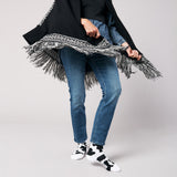 Person wearing a black coat with a fringed scarf, blue jeans, and black and white cow print socks on a light gray background