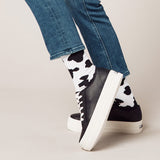 Black sneakers with white soles worn with blue jeans and cow print socks on a neutral background