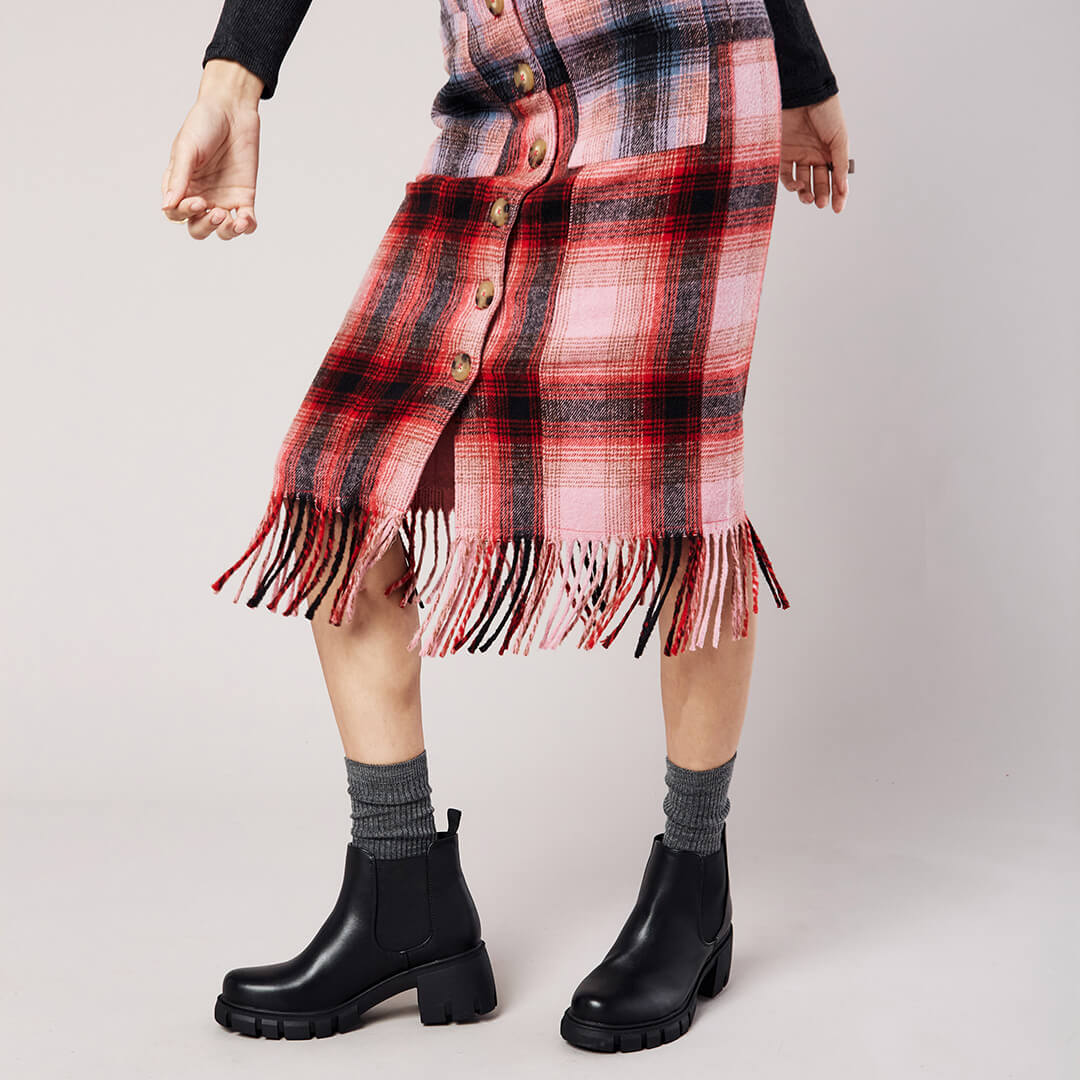 Person wearing a red and black plaid skirt with fringe, black boots, and gray socks on a light background