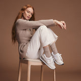 Woman sitting on a stool wearing a beige sweater and socks, white pants, and white sneakers against a brown background thumbnail