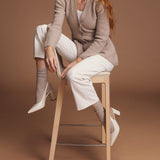 Woman sitting on a stool wearing a beige cardigan and socks, white pants, and high-heeled shoes against a brown background thumbnail