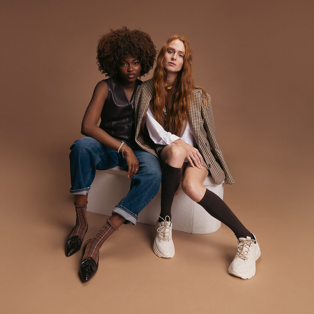 Two women sitting on a white stool against and k.bell socks on a brown background